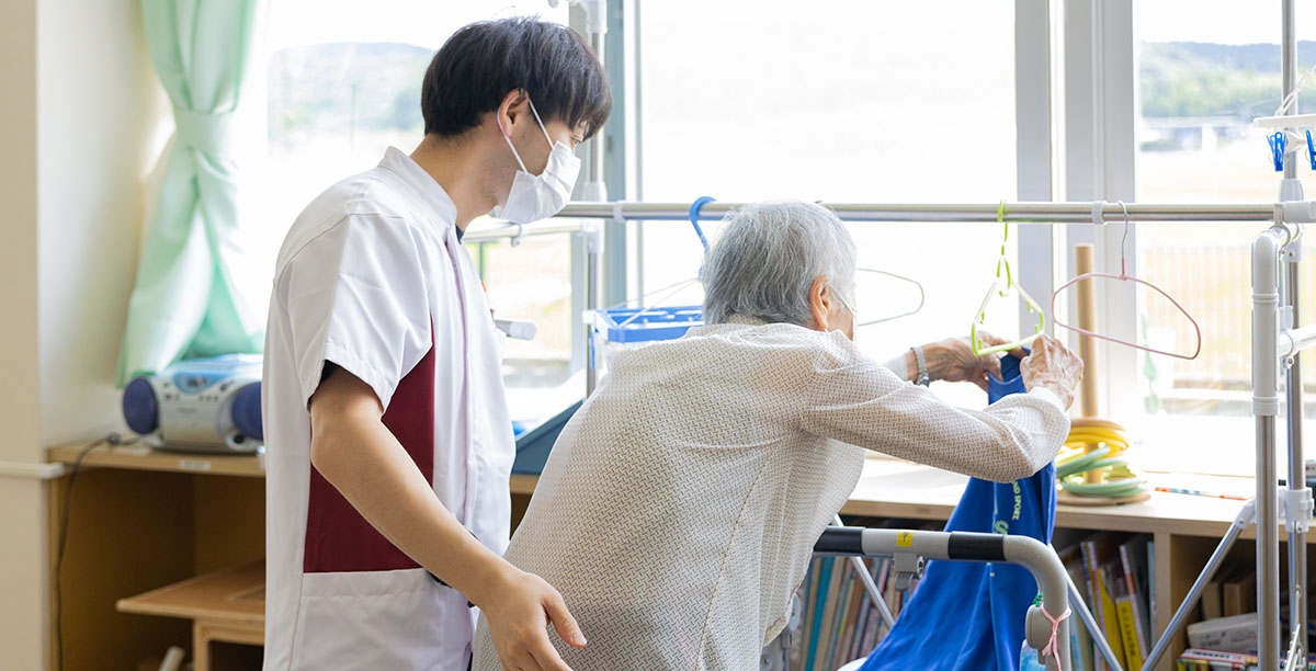 歩行器を使って室内を歩く高齢者と、それを見守る介助者